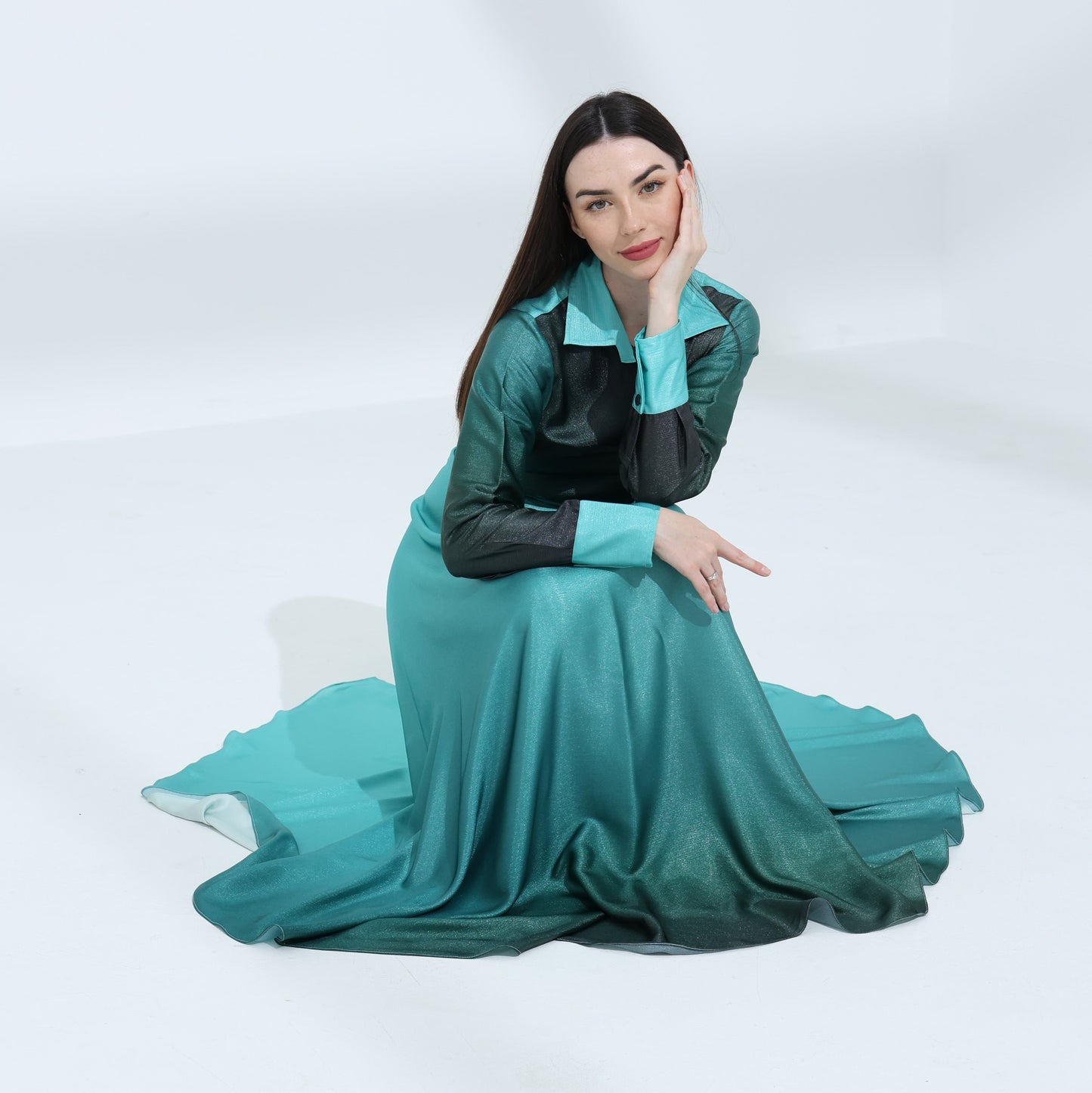 A woman sitting on the floor wearing a long turquoise dress with a solid black top and long sleeves.