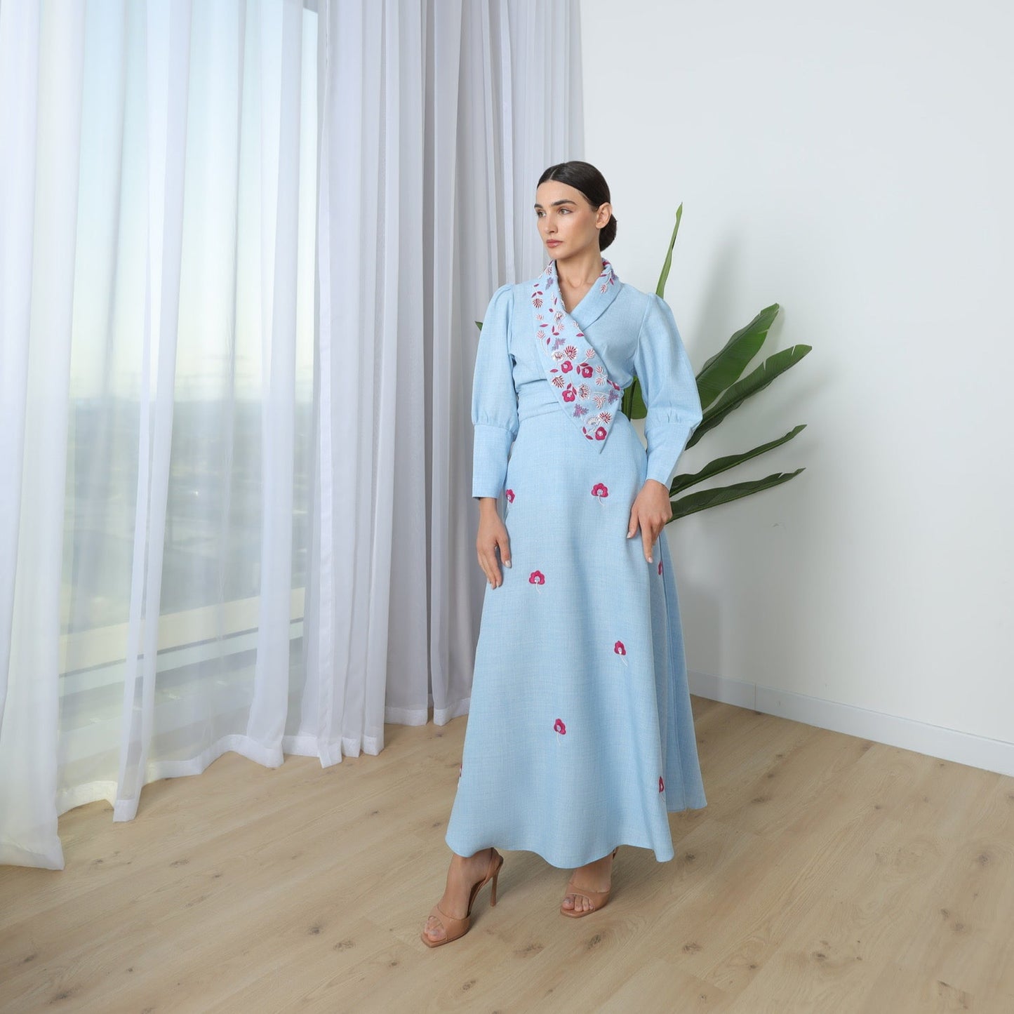 Woman in a light blue dress standing in a room with curtains and a plant.