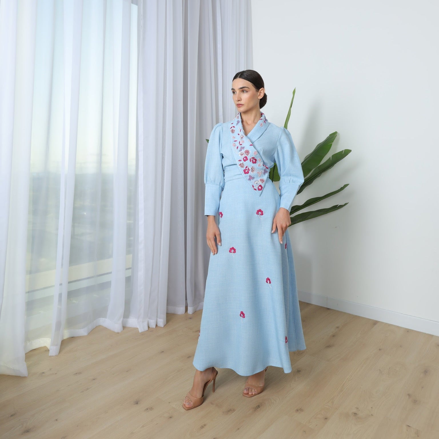 Woman in a light blue dress standing in a room with curtains and a plant.