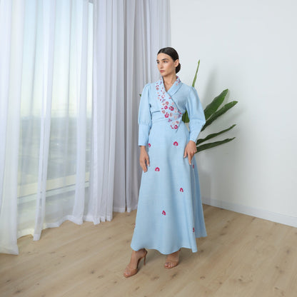 Woman in a light blue dress standing in a room with curtains and a plant.