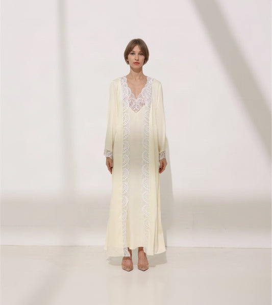 A woman stands in a studio wearing a long cream-colored robe with white lace trim over a matching cream slip dress. The robe has long sleeves and a V-neckline, and the dress has a lace-trimmed neckline and center front panel.